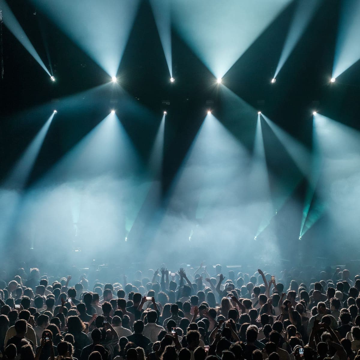 A packed crowd with hands up under teal stage beams.